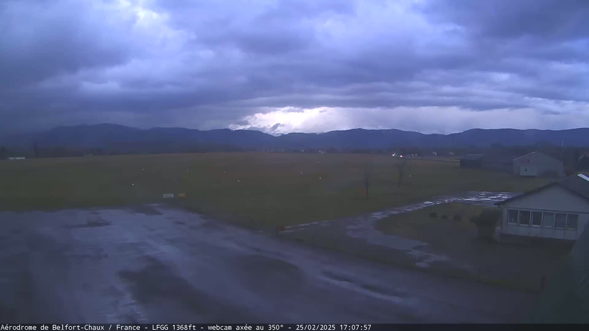A wet airfield sits under a dark, stormy sky before a range of distant mountains.