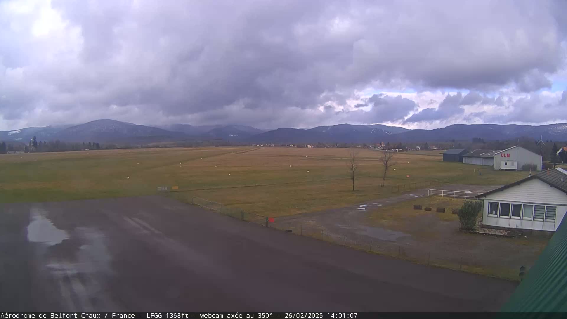 A wet airfield with a hangar and small building is visible, under a cloudy sky, with snow-capped mountains in the background.