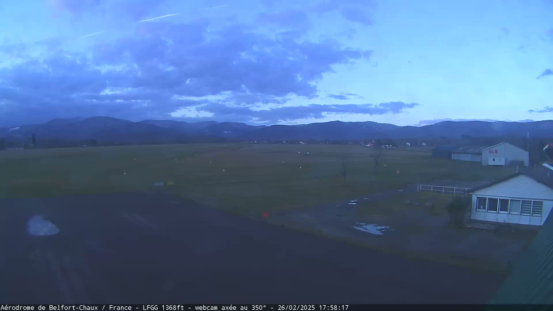 A flat, grassy airfield with a few buildings is visible under a partly cloudy twilight sky, backed by a range of distant mountains.