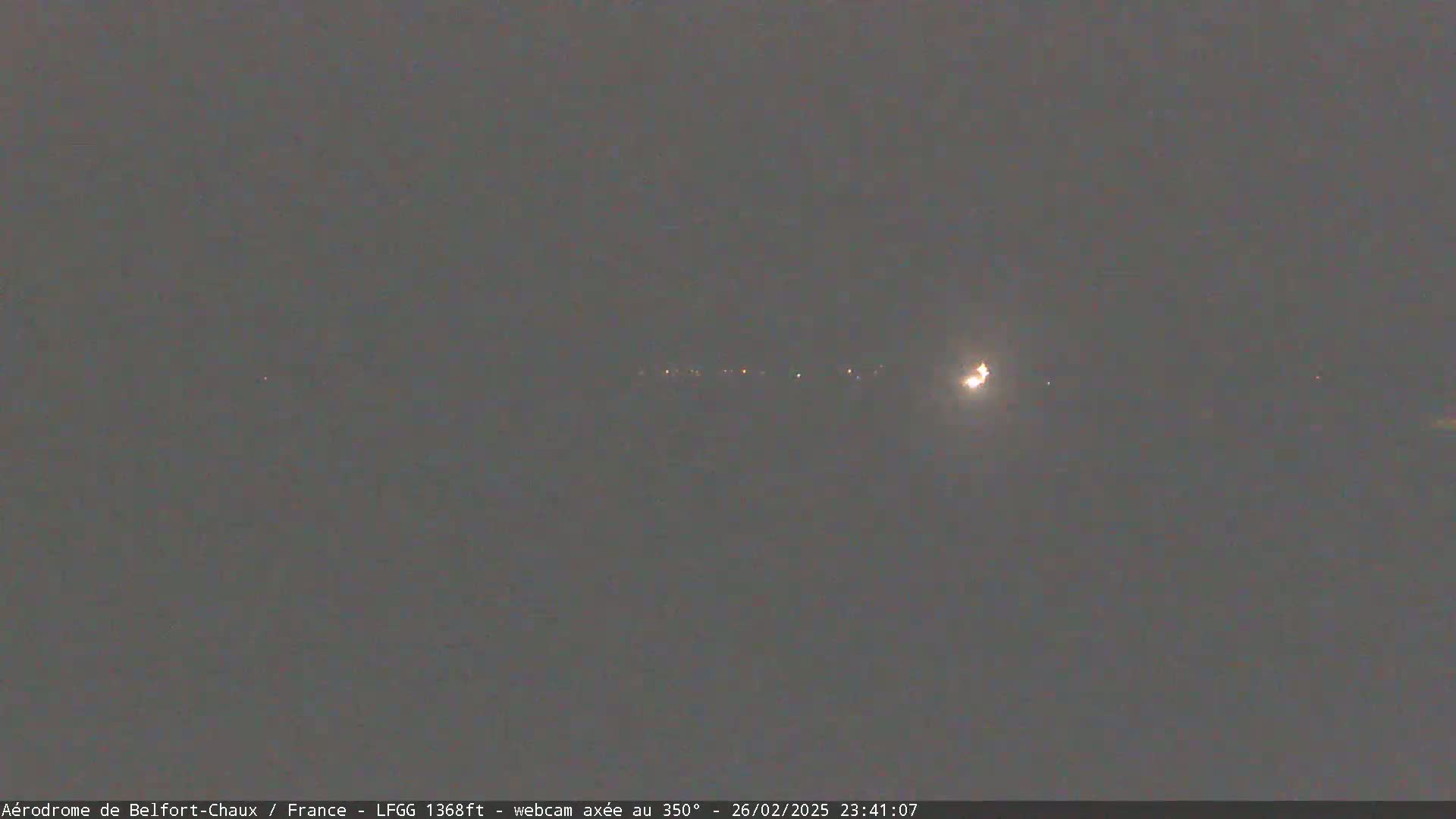 A nighttime long shot shows a line of distant lights and a brighter light, likely a plane, in a dark, overcast sky.