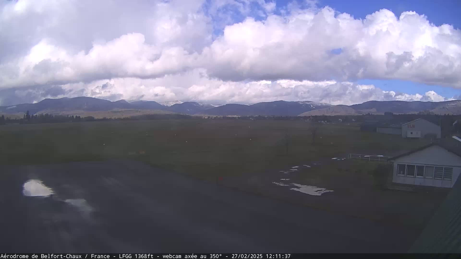 A partly cloudy day reveals a flat, grassy field with a small building and a paved area, all set against a backdrop of distant, snow-capped mountains.