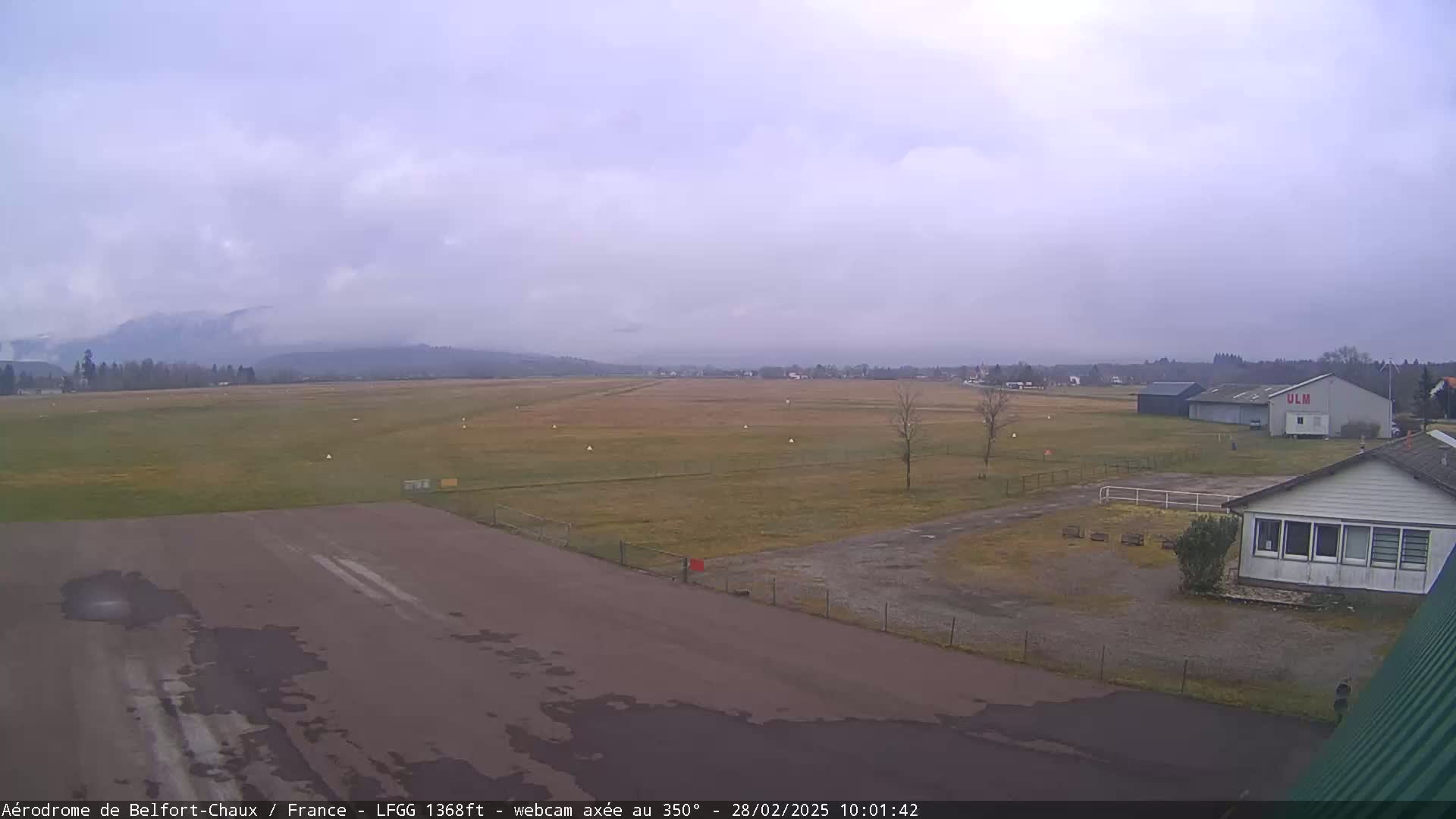 A mostly cloudy day reveals a flat airfield with a runway, small buildings, and distant, fog-covered mountains.