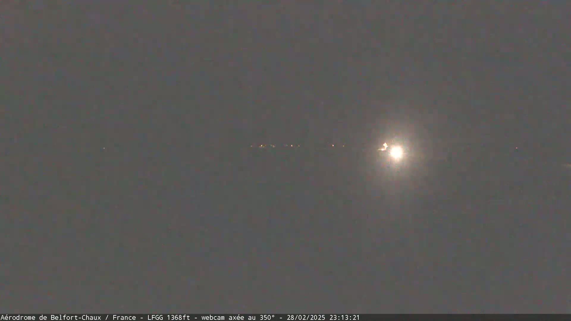 A bright light, possibly the moon, shines in a night sky over a distant line of faint lights.