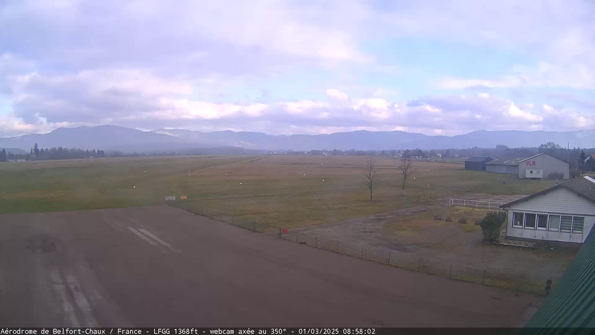 A mostly cloudy day reveals a small airfield with a runway, several buildings, and a distant mountain range.