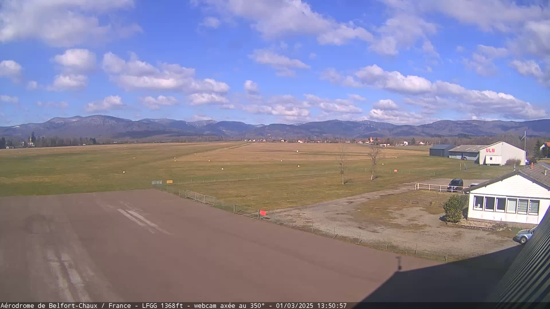 A mostly sunny day shows a small airfield with a runway, buildings, and vehicles, all set against a backdrop of distant mountains.