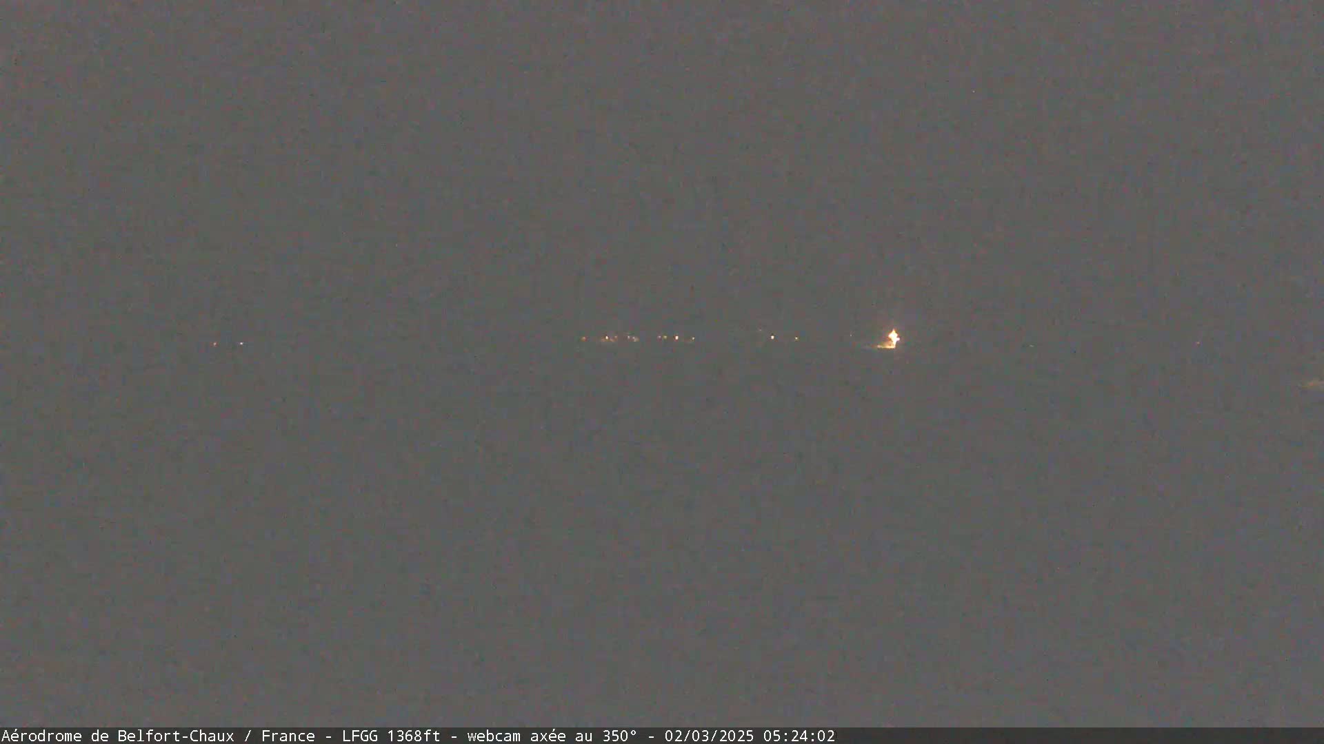 A dimly lit, nighttime landscape shows a line of faint lights in the distance under a dark, overcast sky.