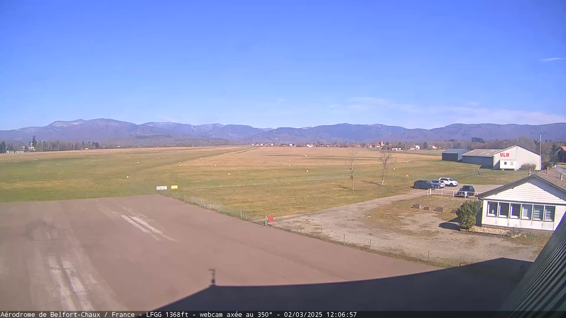 A small airfield with a runway, several buildings, and a few cars is situated in a grassy field, with distant mountains visible under a clear blue sky.