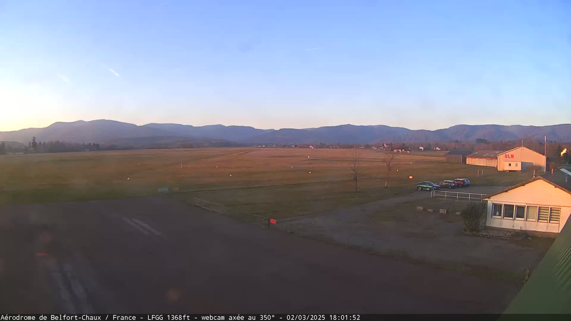 A flat, grassy airfield with a few parked cars and buildings is situated in front of a range of distant mountains under a clear, twilight sky.