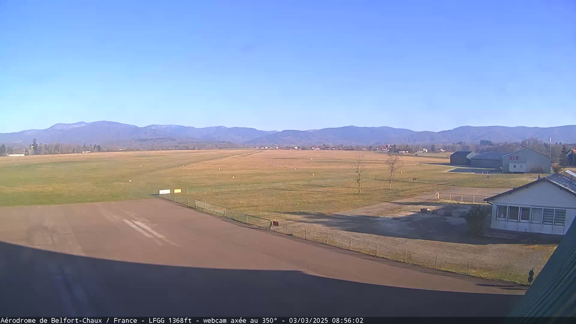 A clear, sunny day at a small airfield with a runway, hangar, and surrounding buildings, set against a backdrop of distant mountains.