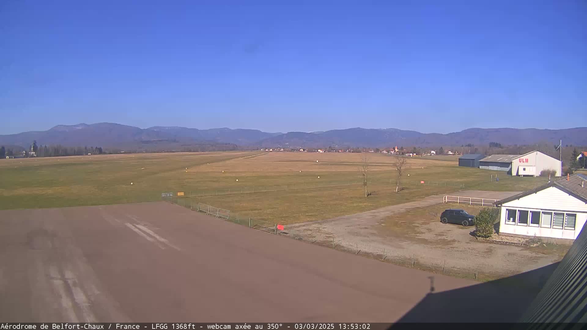 A small airfield with a runway, a few buildings, and a car is shown on a sunny day with mountains in the background.