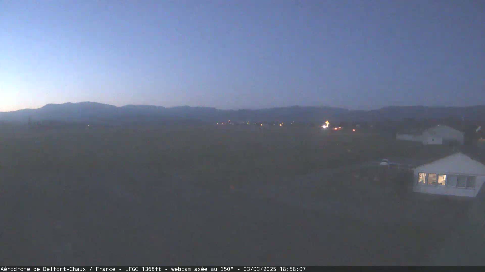 A hazy, twilight view shows a flat, dark landscape with a few buildings and distant mountains under a dusky blue sky.