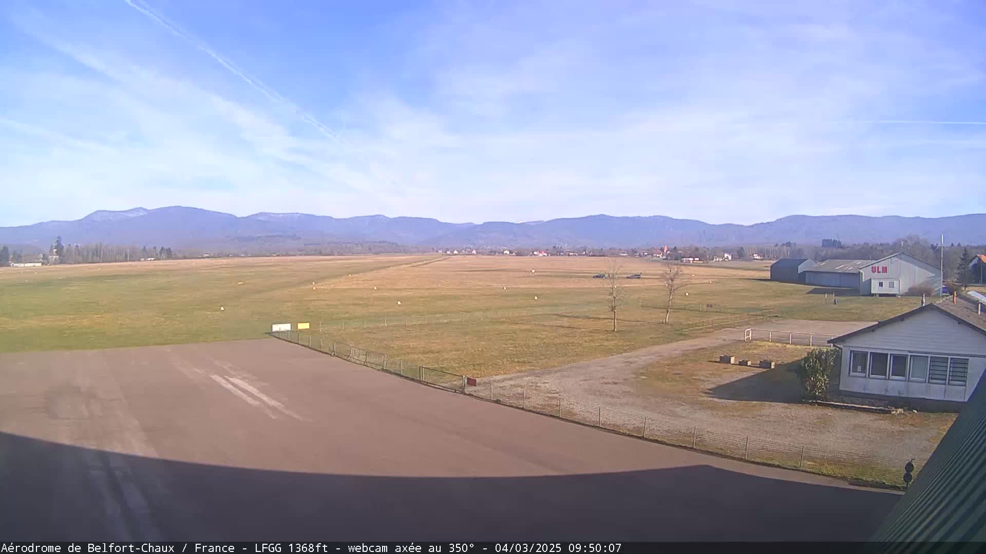 A mostly sunny day at an airfield shows a runway leading to a hangar and other buildings, with distant mountains in the background.