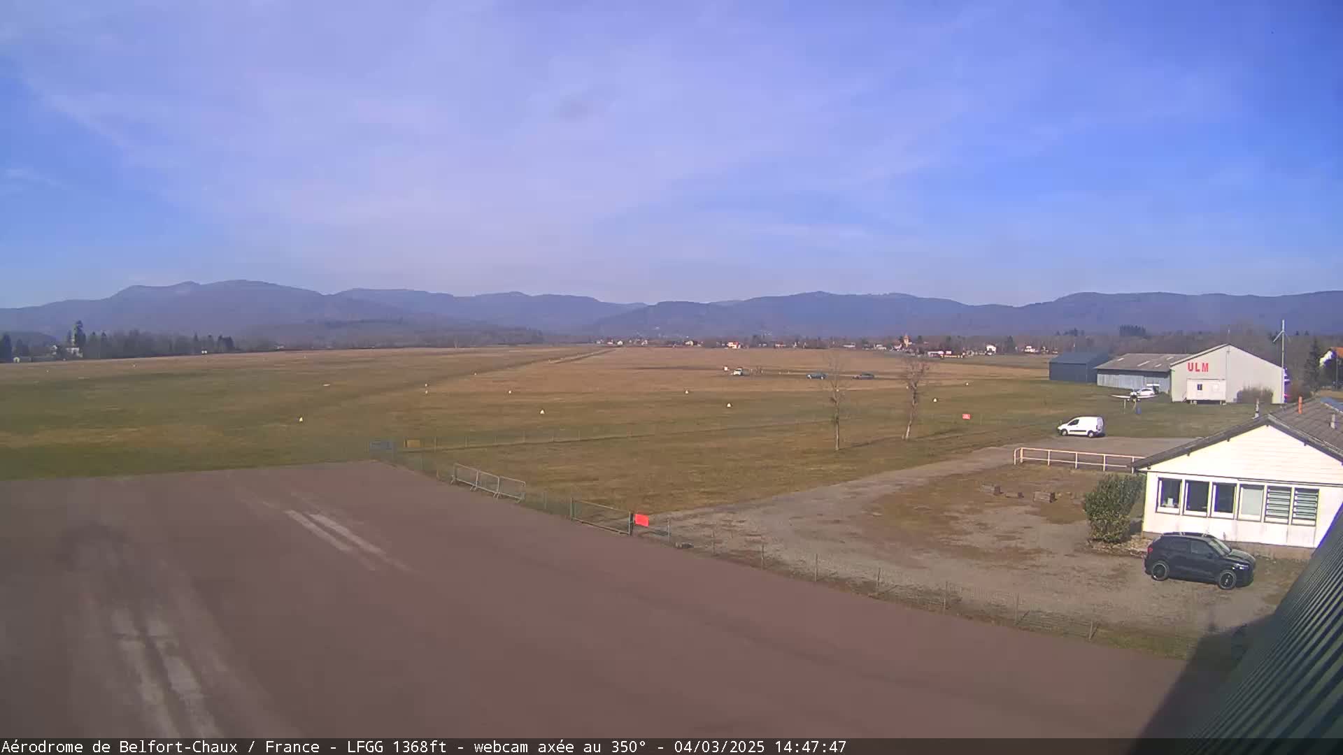 A small airfield with a hangar, a few vehicles, and a small building is situated in a grassy field, with mountains in the background under a clear blue sky.