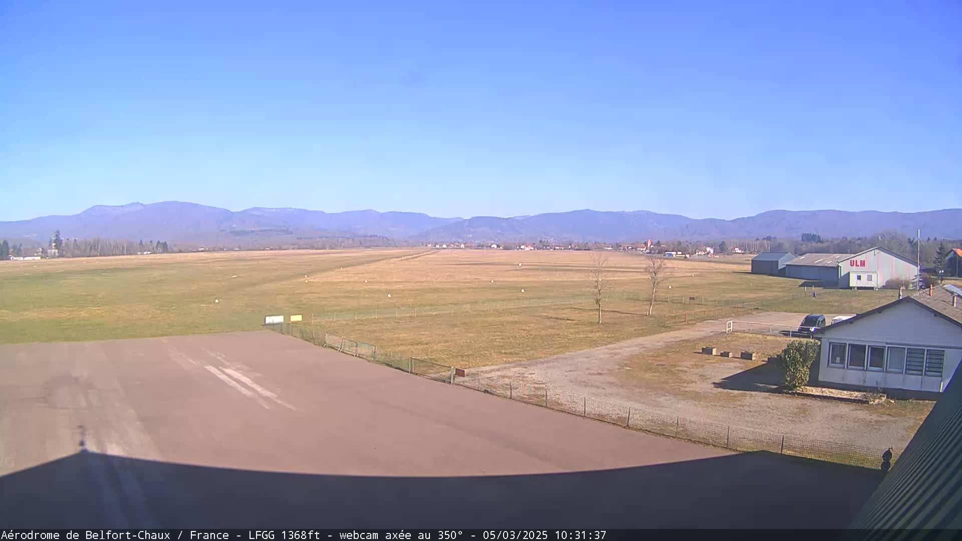 A long airfield runway stretches toward distant mountains under a clear blue sky.