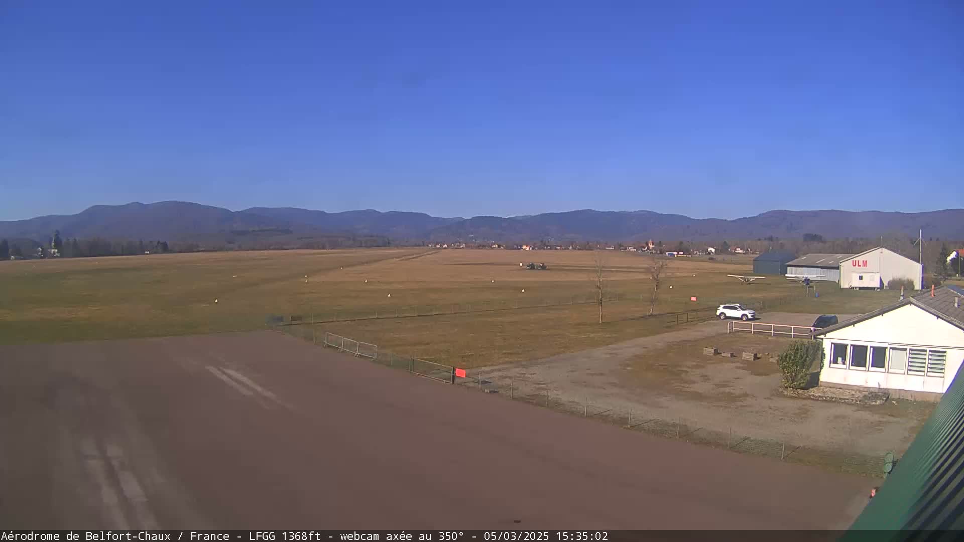 A small airport with a runway, a few small planes, and buildings sits before a range of distant mountains under a clear blue sky.