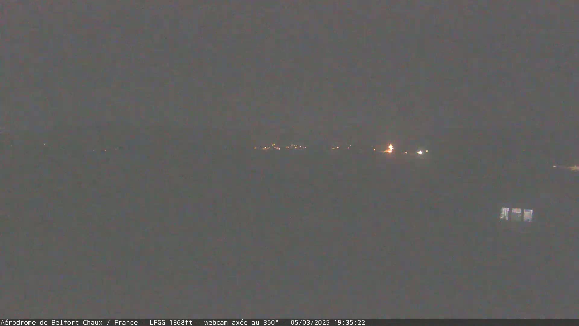 A dimly lit, nighttime landscape shows a few faint lights in the distance under a dark, overcast sky.