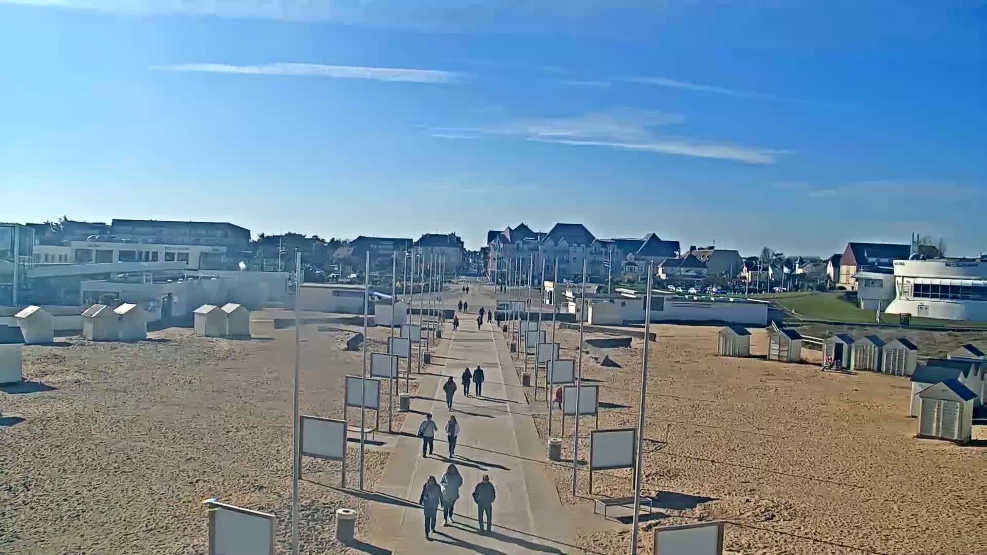 A paved walkway lined with blank signs leads from a sandy beach toward buildings and a clear blue sky.