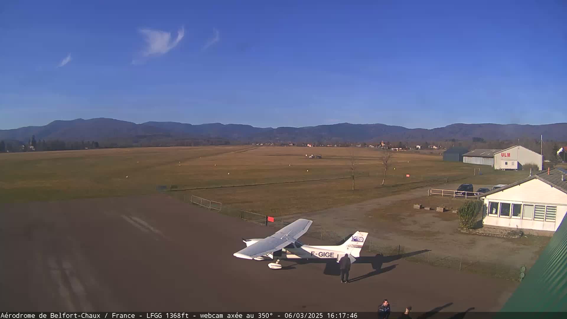 A small airplane sits on a tarmac at a small airport, with distant mountains visible under a clear blue sky.