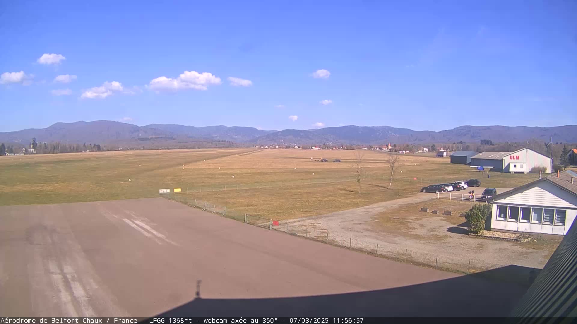 A small airfield with a runway, several buildings, and a few parked cars is seen on a sunny day with few clouds, set against a backdrop of distant mountains.