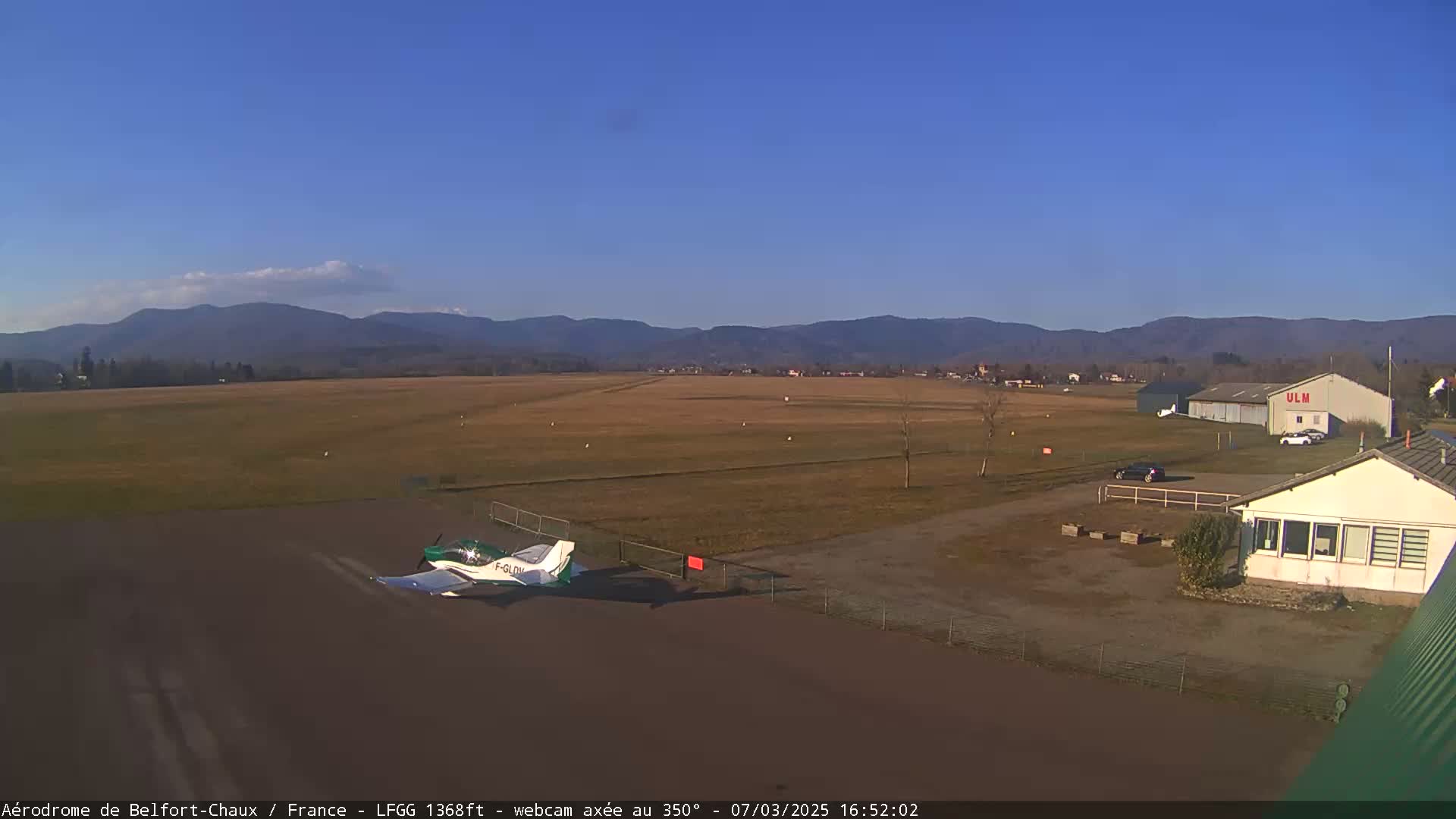 A small green airplane sits on a tarmac at a small airfield under a clear blue sky, with distant mountains in the background.