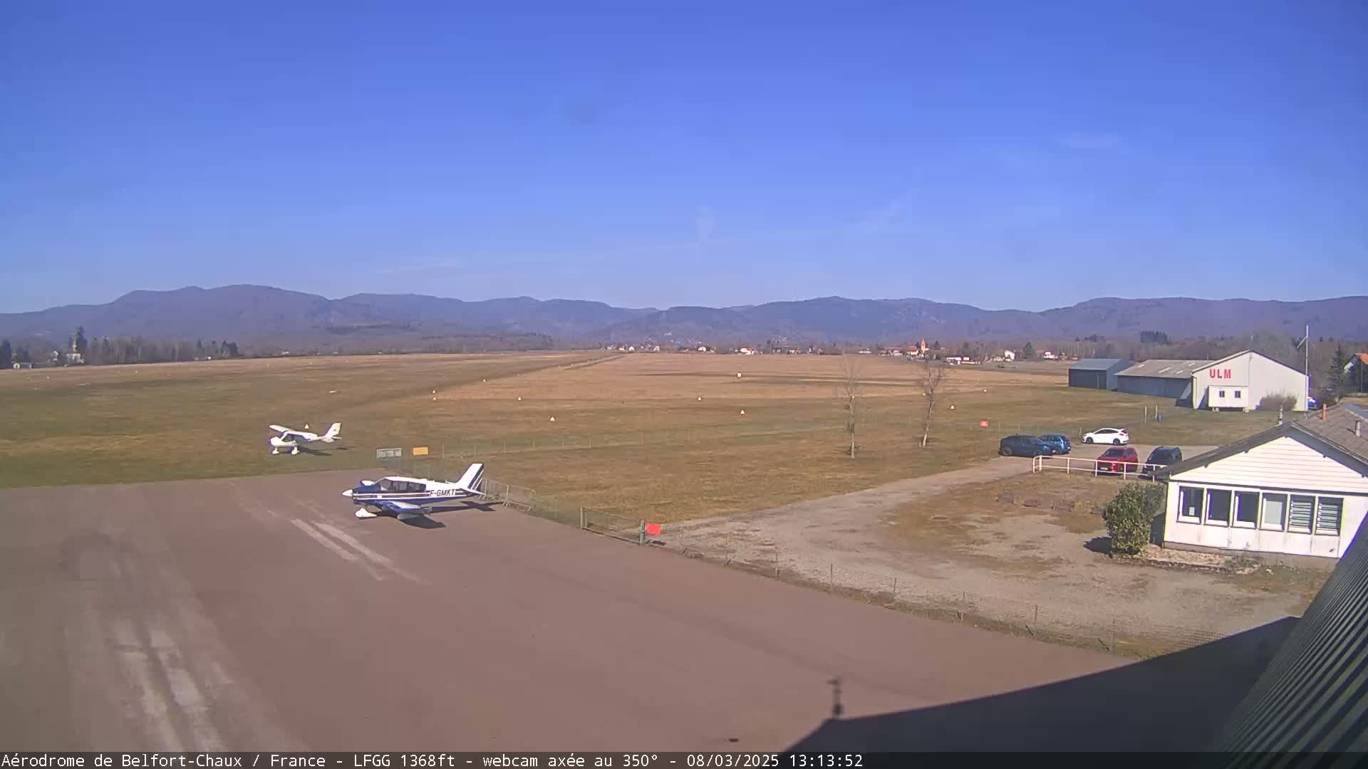 Two small airplanes are parked on a tarmac at a small airport on a clear, sunny day, with distant mountains visible in the background.