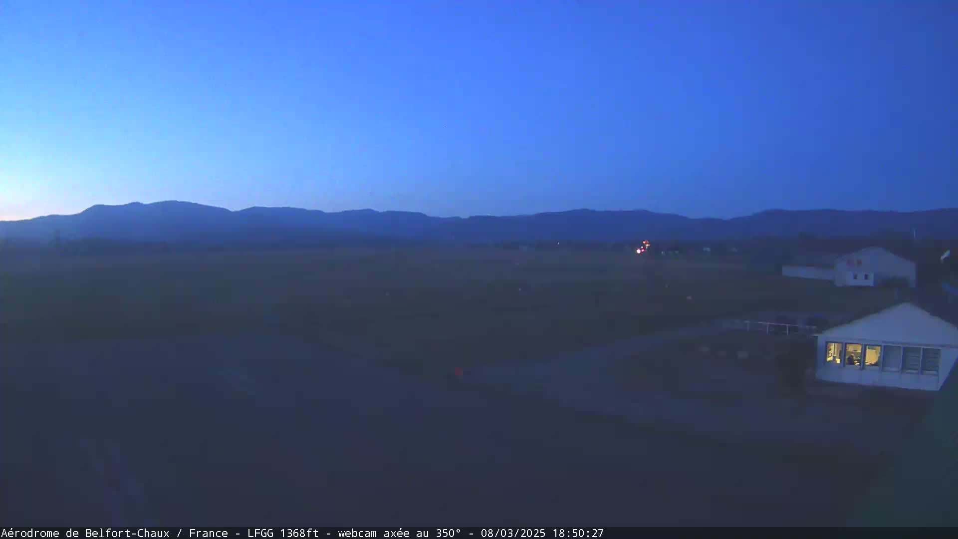 A twilight view shows a flat landscape with distant mountains under a clear, dark blue sky, a few buildings are visible in the foreground.