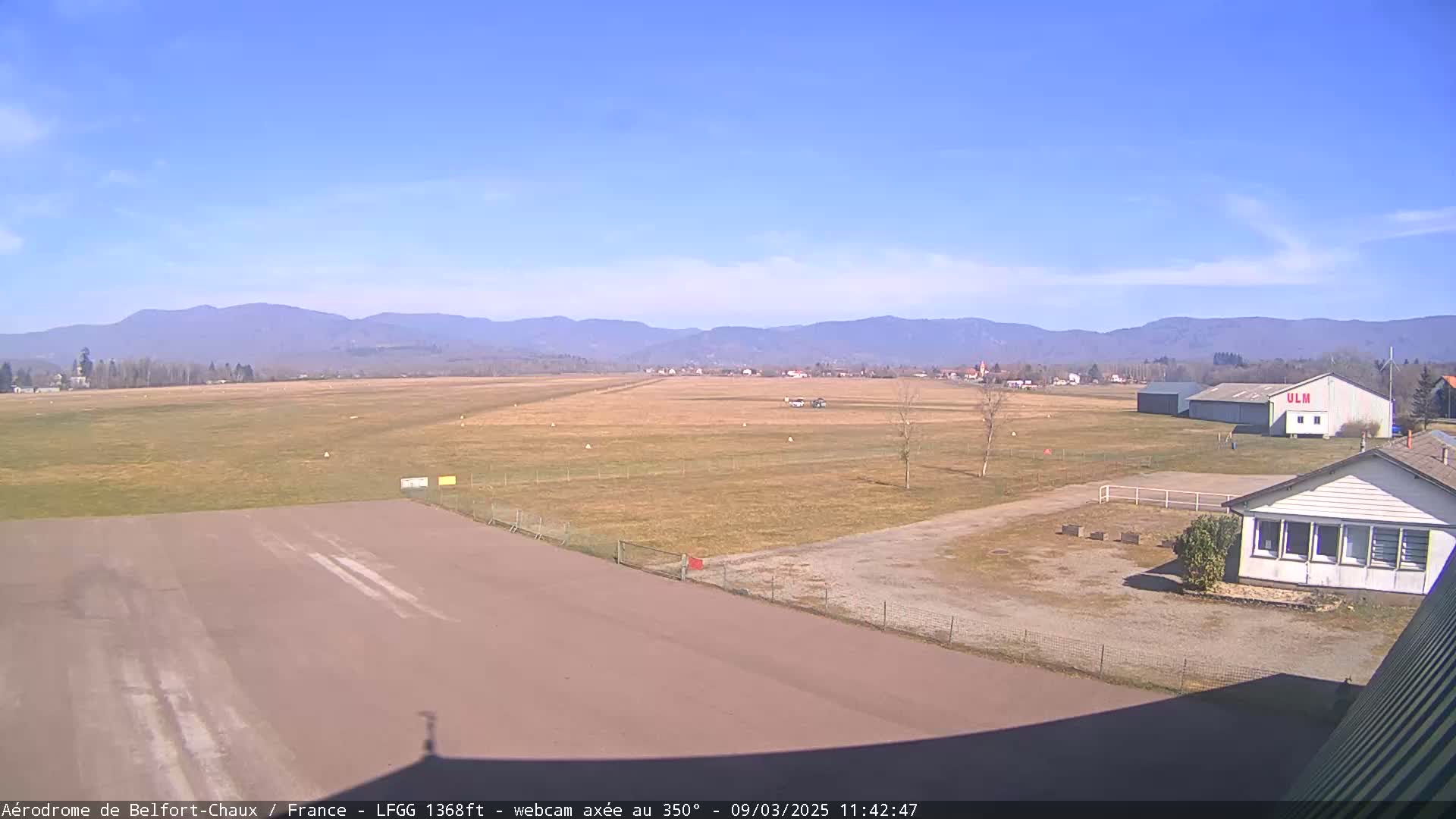 A clear, sunny day at an airfield shows a runway and surrounding buildings with distant mountains in the background.