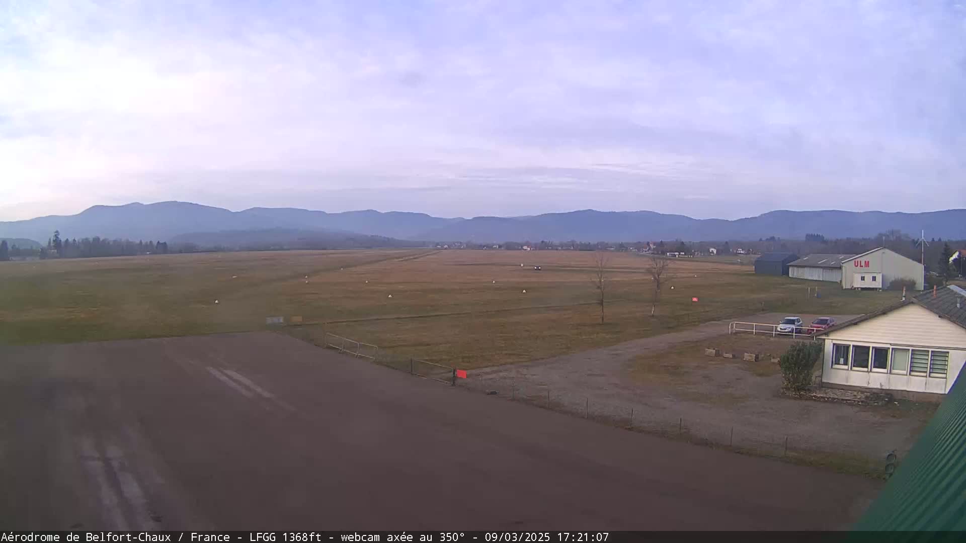A grassy airfield with a runway, a few buildings, and distant mountains under a partly cloudy sky.