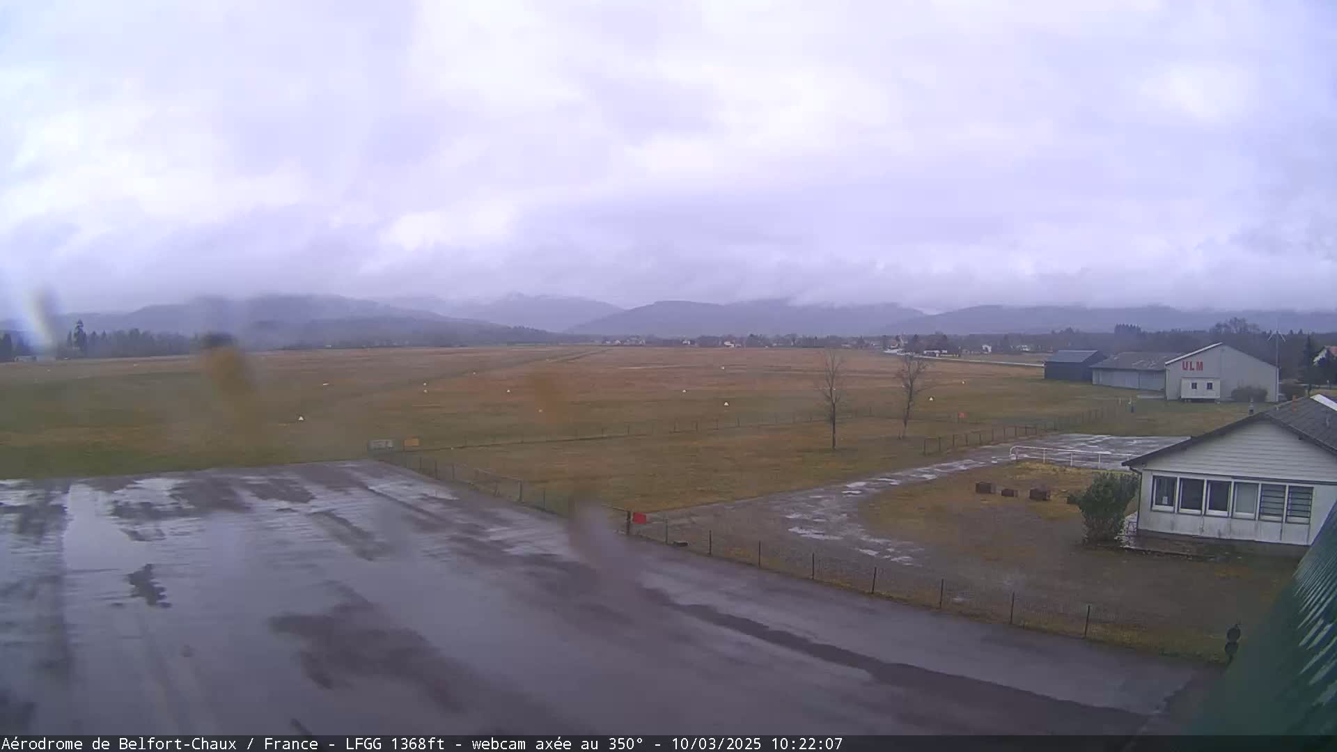 A wet, overcast day at a small airfield with a few buildings, surrounded by a grassy field and distant mountains.
