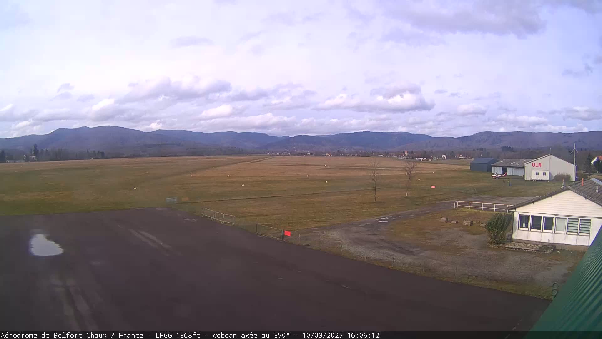 A mostly clear day at an airfield shows a runway, several buildings, a small plane, and distant mountains.