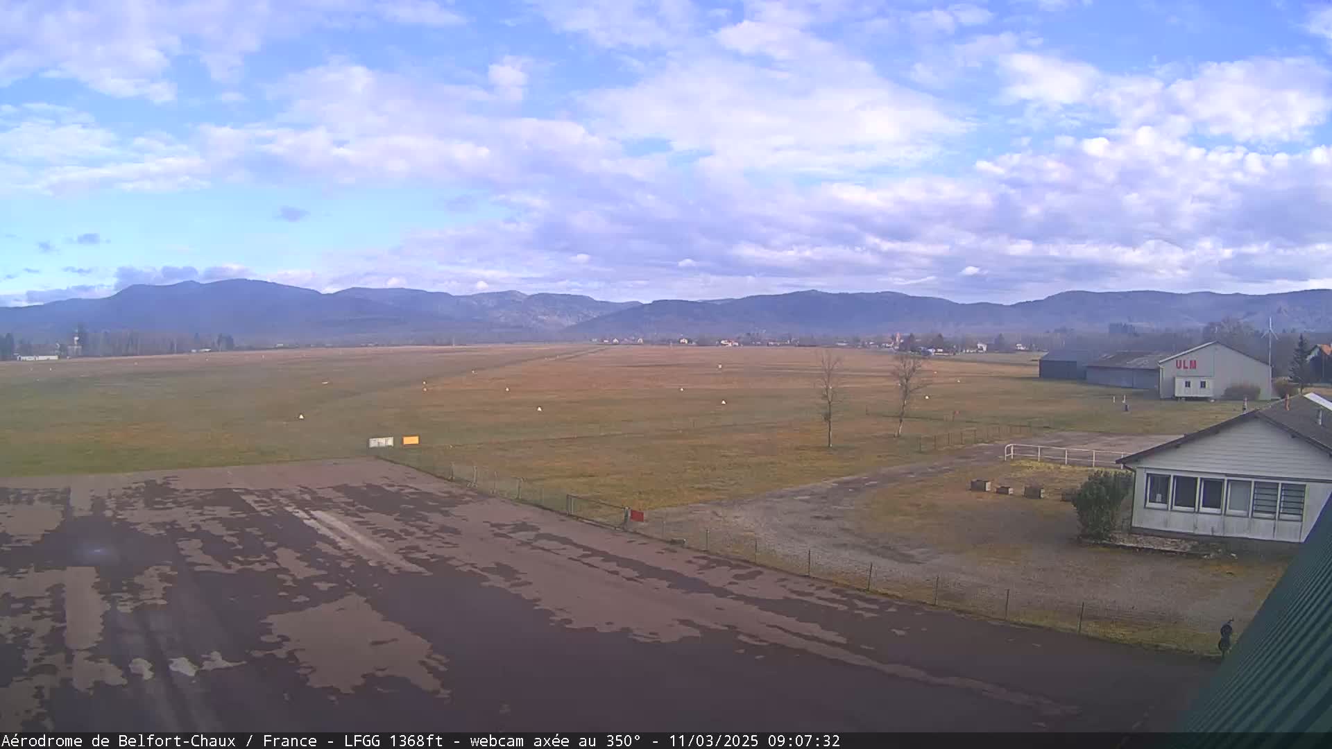 A mostly clear day with some clouds over a flat, grassy airfield next to a small building, with distant mountains in the background.