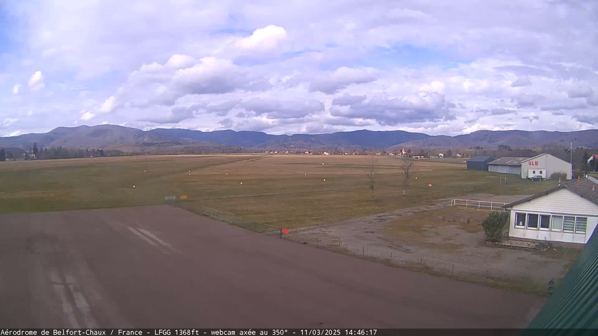 A mostly cloudy day reveals a small airfield with a runway, buildings, and distant mountains.