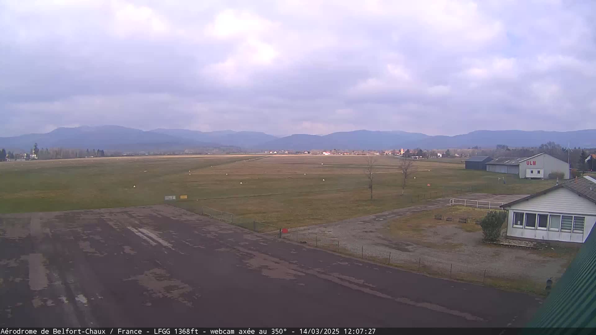 A small airfield with a runway and buildings sits before a range of distant mountains under a mostly cloudy sky.