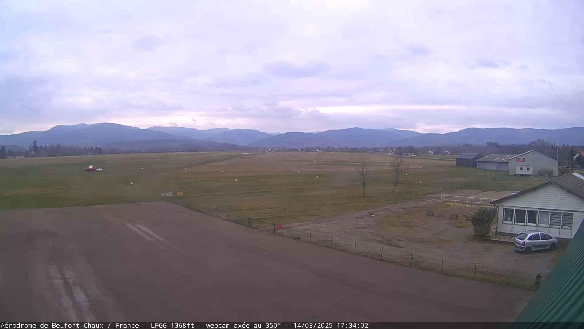 A small airfield with a runway and a few buildings is situated in a flat grassy area, overlooked by distant mountains under an overcast sky.