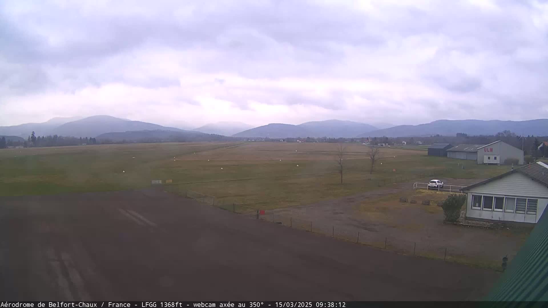 A small airfield with a few buildings and a car is visible, set against a backdrop of distant mountains under an overcast sky.
