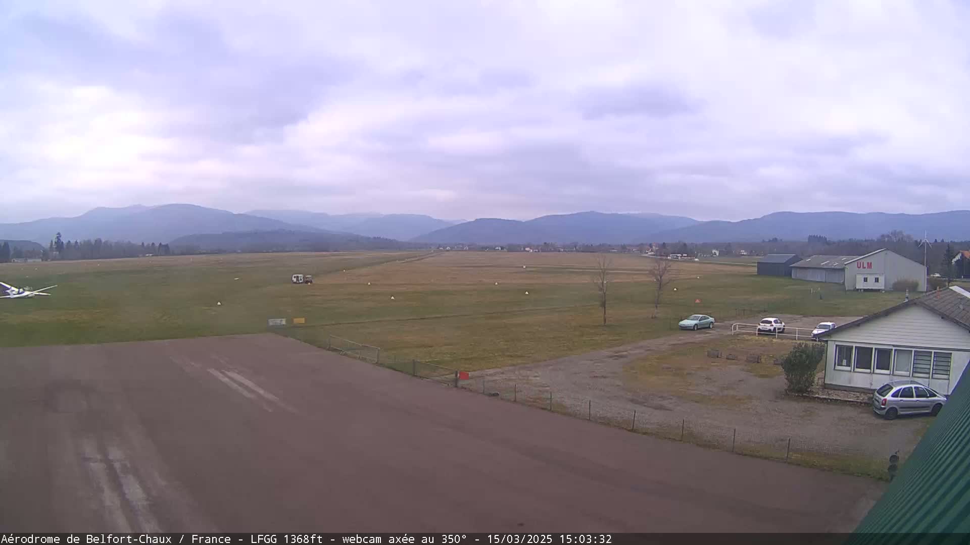 A small airfield with several cars and a small plane on an overcast day, with distant mountains visible in the background.