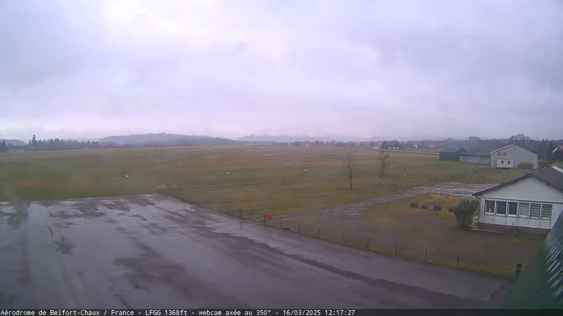 A wet airfield with a building and grassy areas is visible under an overcast sky.