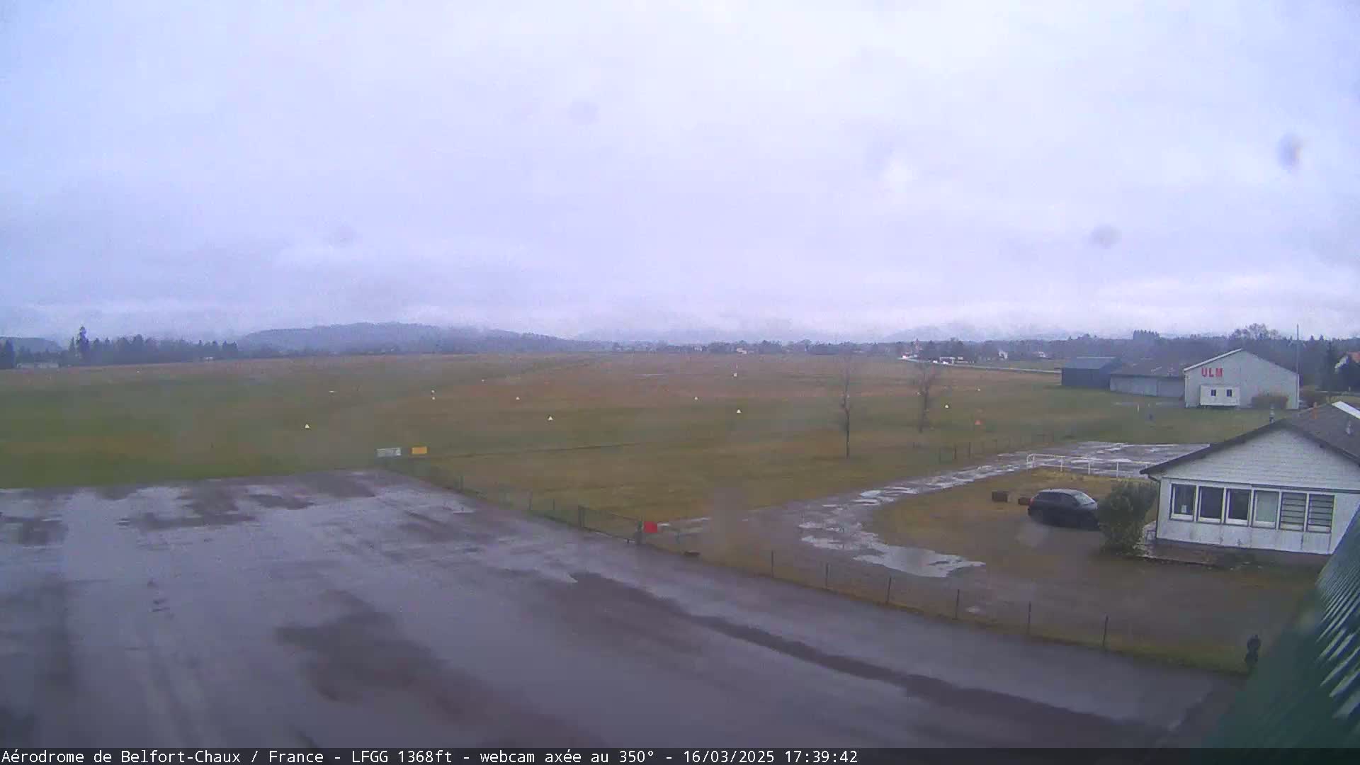 A wet airfield with a small building and a car is visible under an overcast sky.