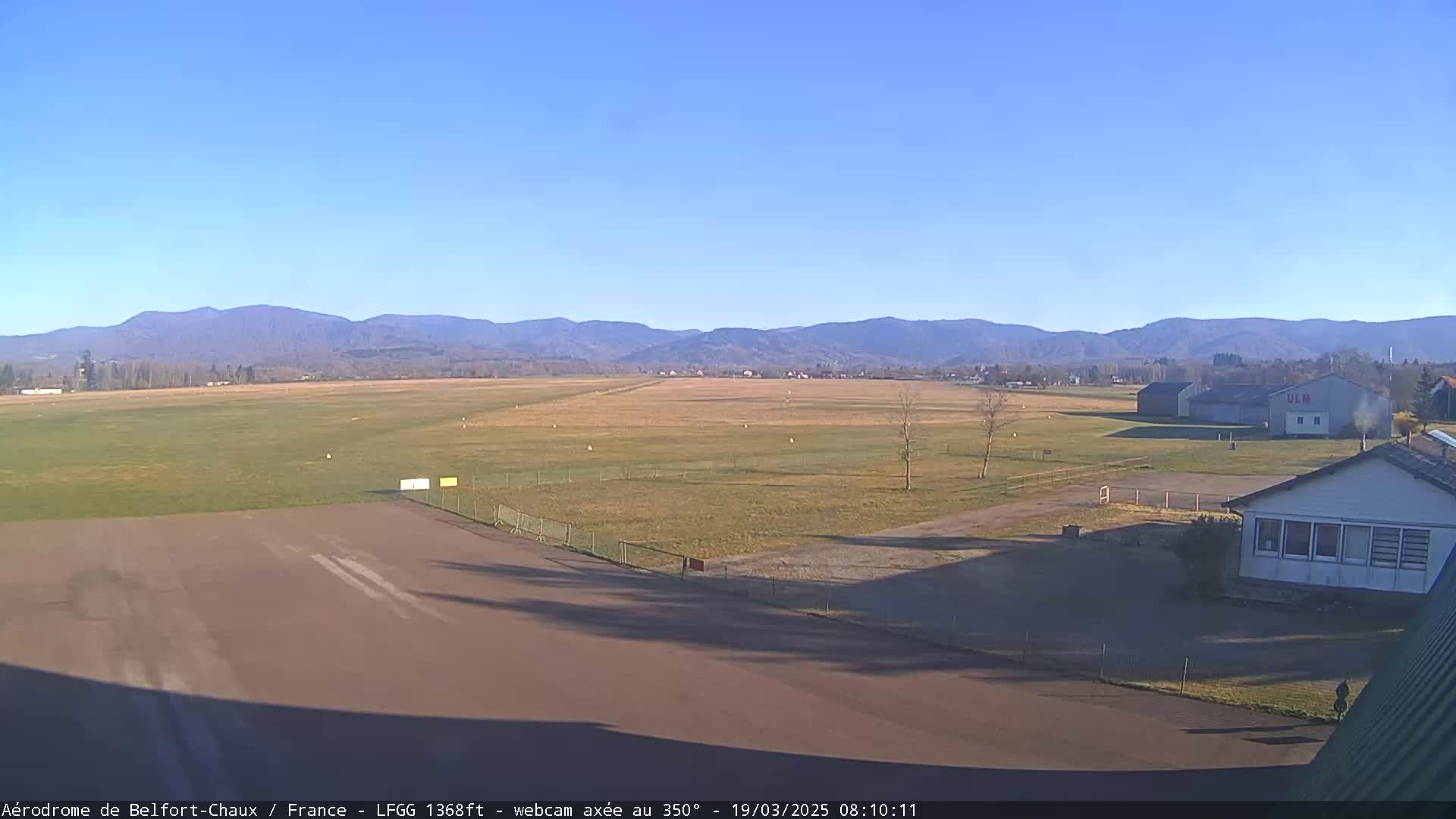 A grassy airfield with a runway and several buildings is visible beneath a clear blue sky and distant mountains.