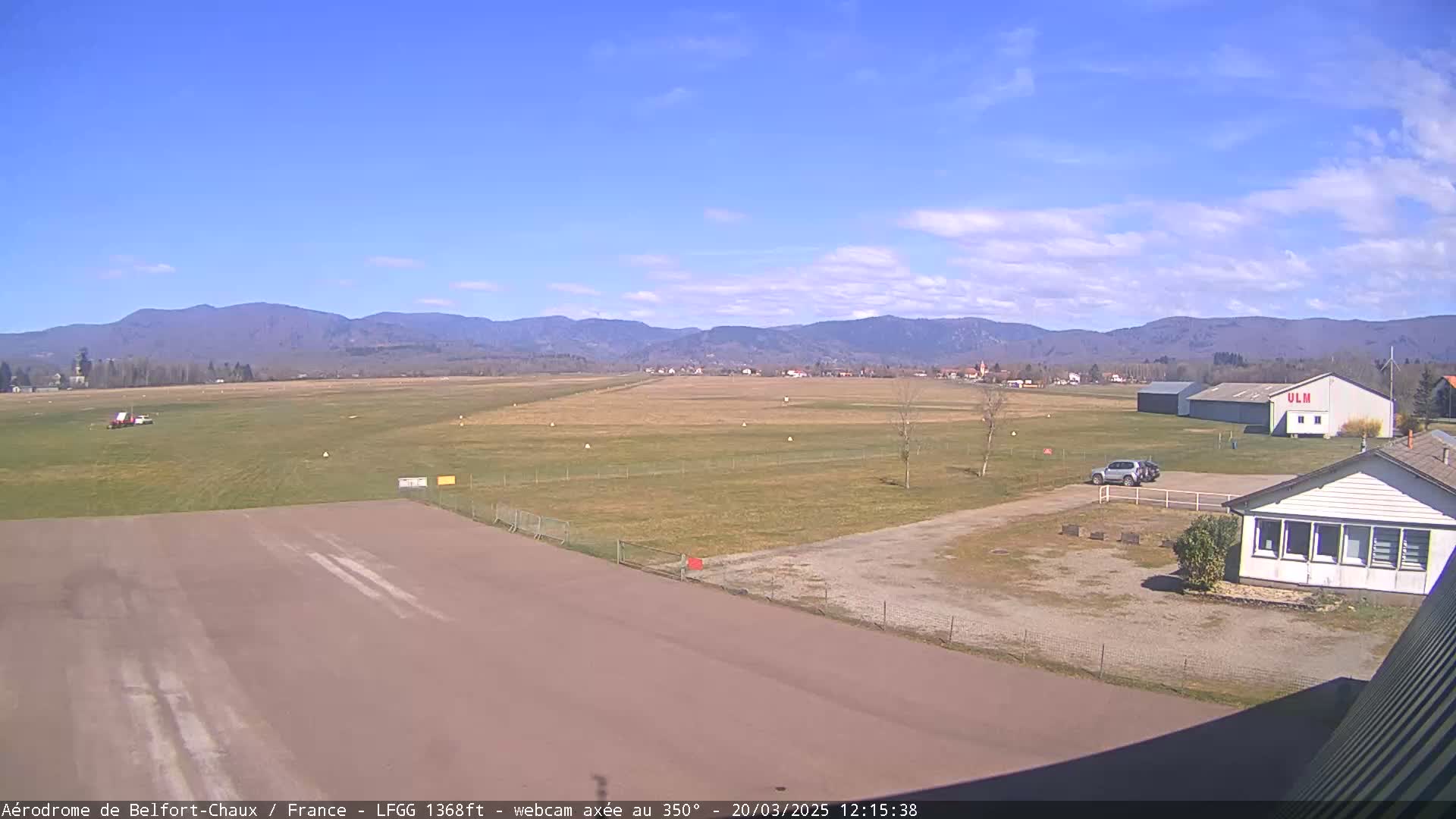 A mostly sunny day at an airfield, showing a runway, a few buildings, and distant mountains.