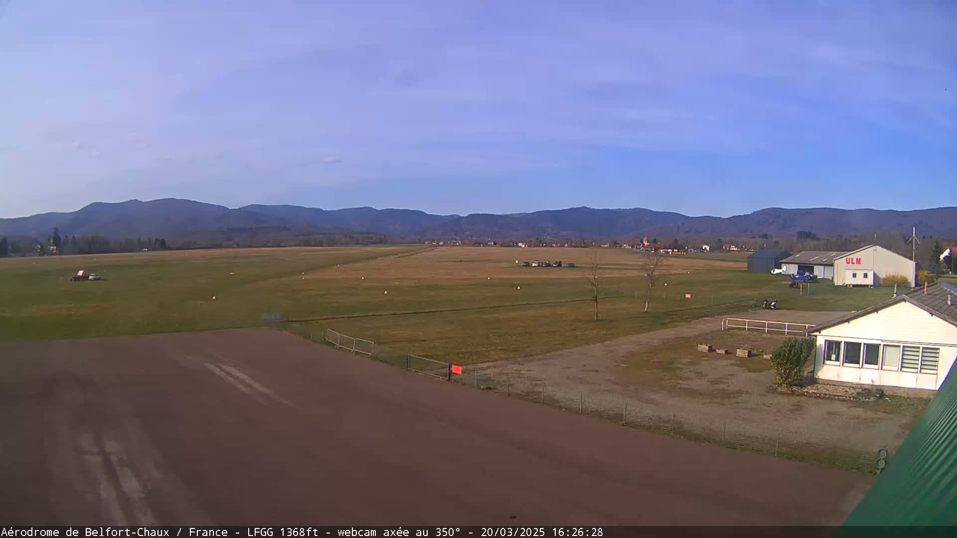 A small airfield with a runway, a few buildings, and a small aircraft, situated in a grassy field against a backdrop of distant mountains under a clear, bright sky.