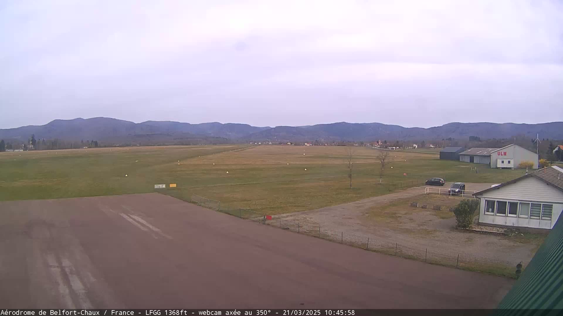 A small airfield with a runway and several buildings sits before a range of distant, low mountains under a mostly cloudy sky.