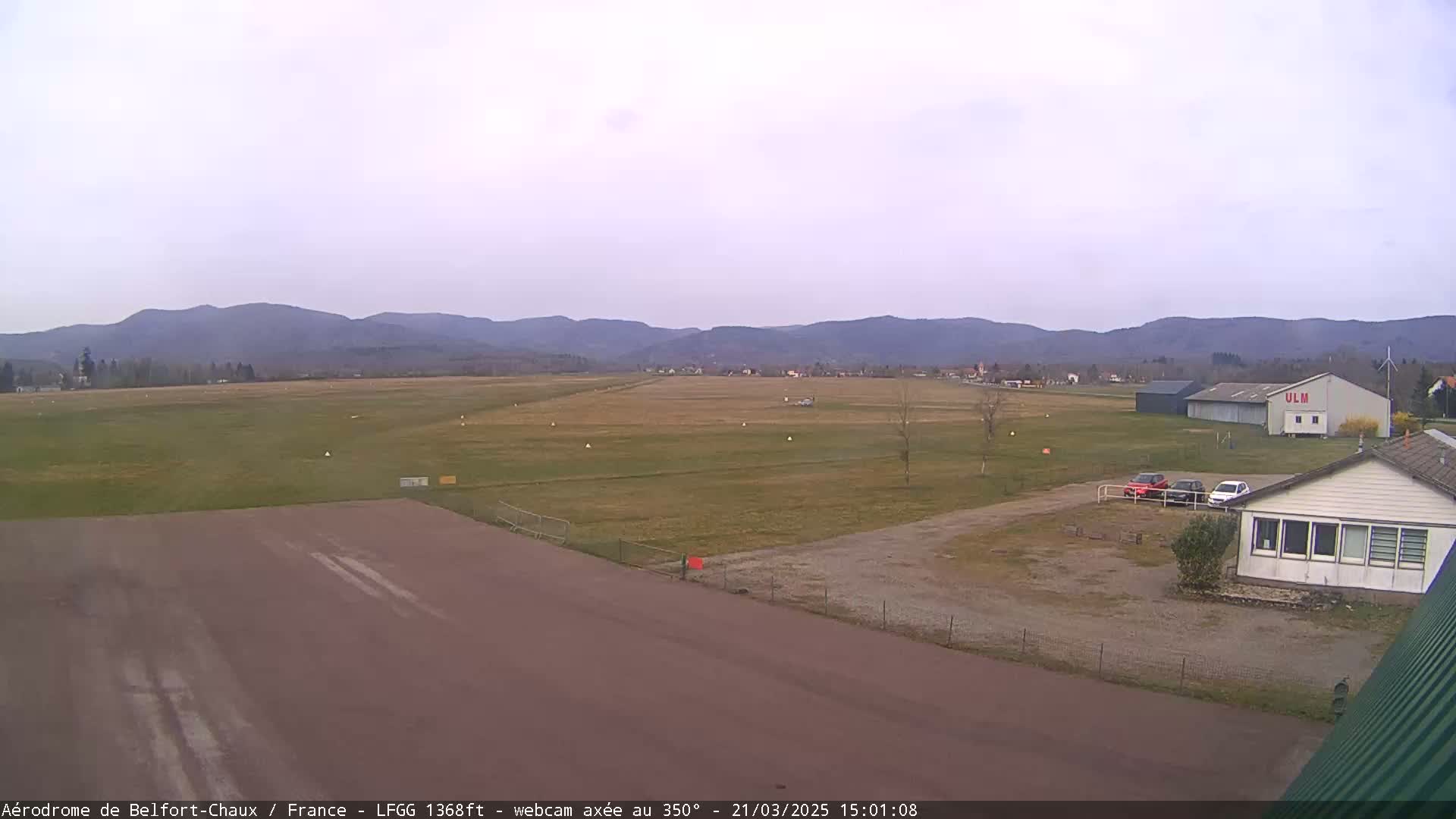 A small airfield with a runway, a few buildings, and several cars is situated in front of a mountain range under an overcast sky.