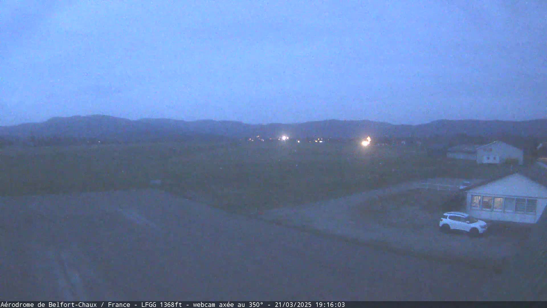 A twilight view of a flat, sparsely populated landscape with a few houses and a single car visible, and distant hills under a clear, dark blue sky.