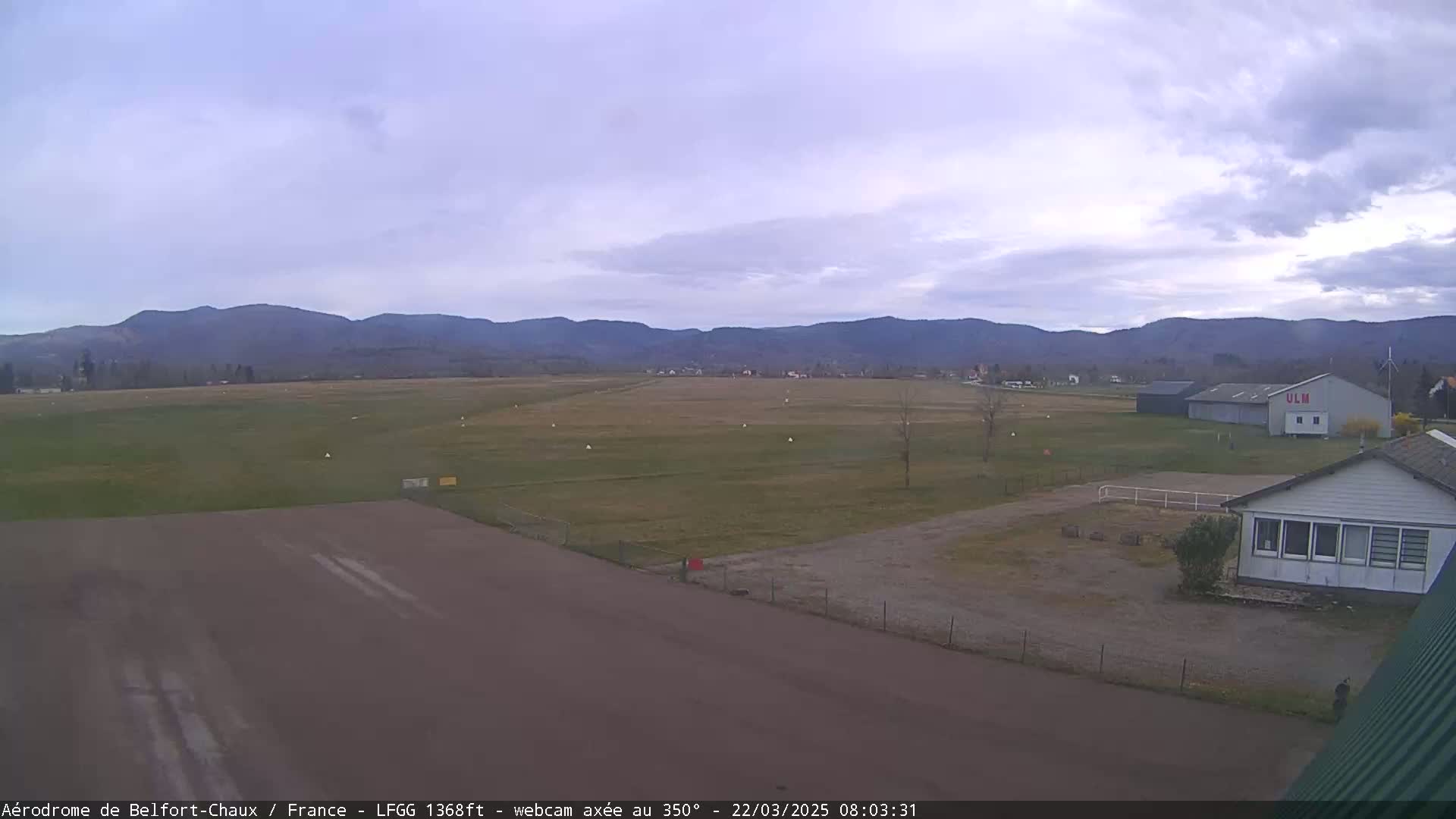 A mostly cloudy day reveals a flat, grassy airfield bordered by a small building and distant mountains.