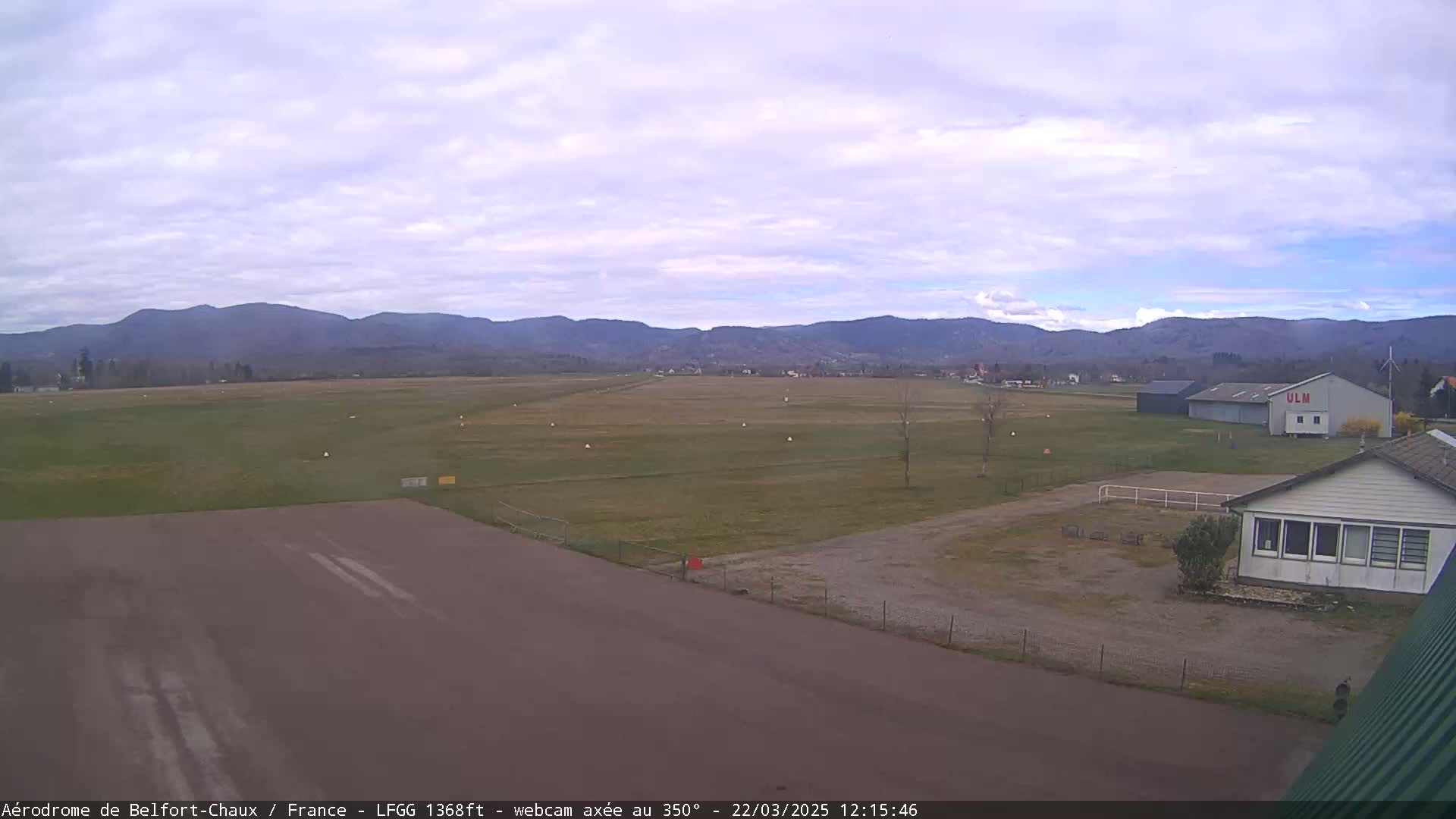 A mostly cloudy day reveals a view of a grassy airfield with a runway, hangar, and small building, all set against a backdrop of distant mountains.