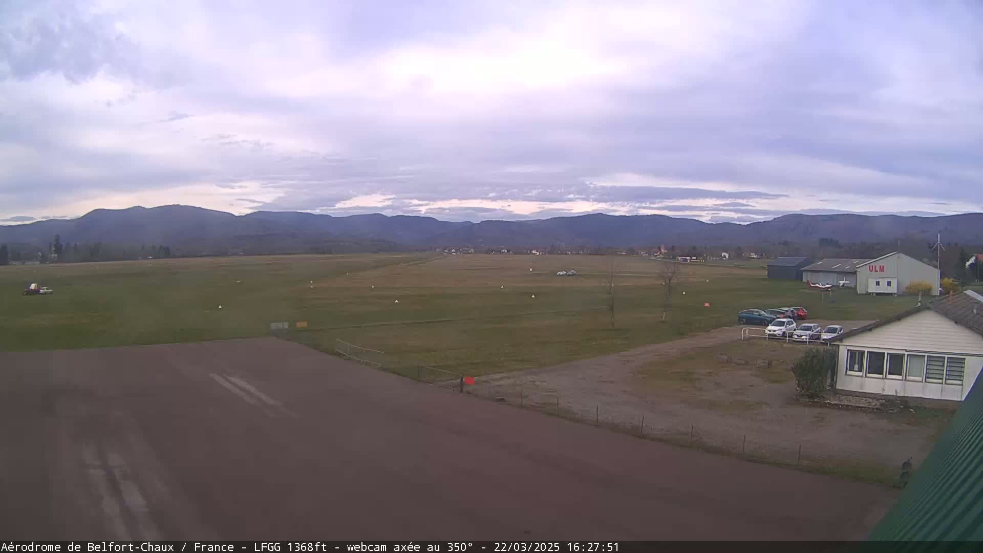 A mostly clear, overcast day reveals a small airfield with several parked cars and a hangar, backed by distant mountains.