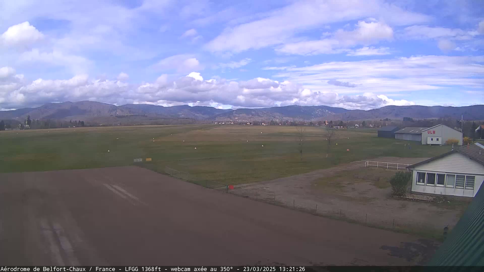 A mostly sunny airfield with a runway, grassy fields, and a few buildings is situated against a backdrop of distant mountains under a partly cloudy sky.