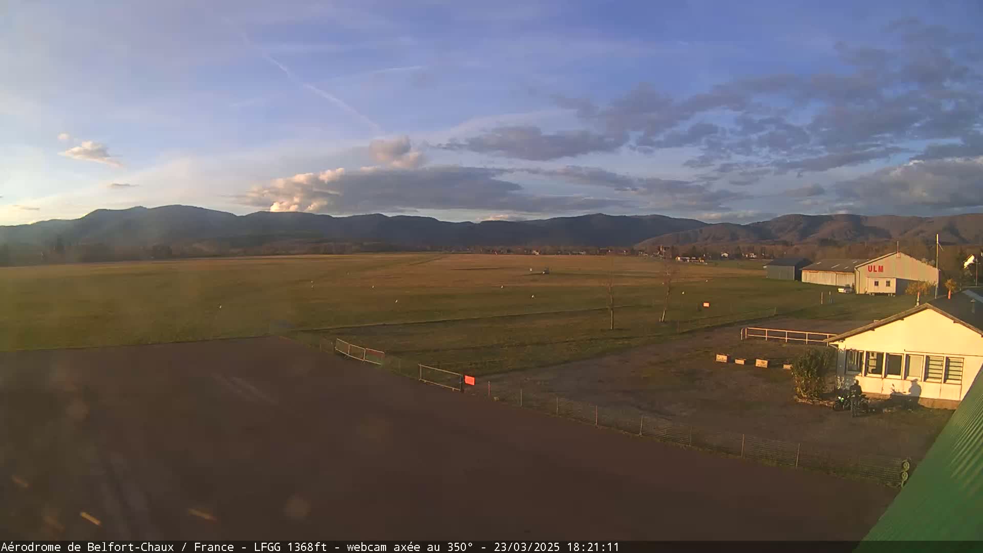 A mostly clear sky over a flat, grassy airfield with a few buildings and distant mountains.