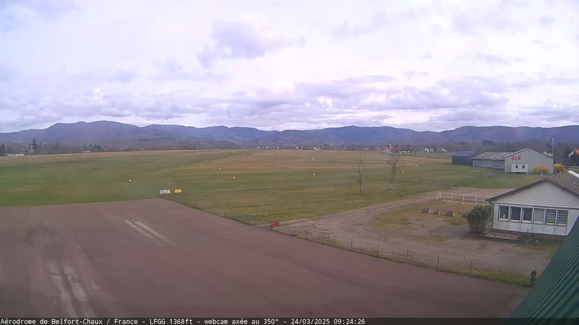A mostly cloudy day reveals a grassy airfield with a runway and buildings, backed by a range of distant mountains.